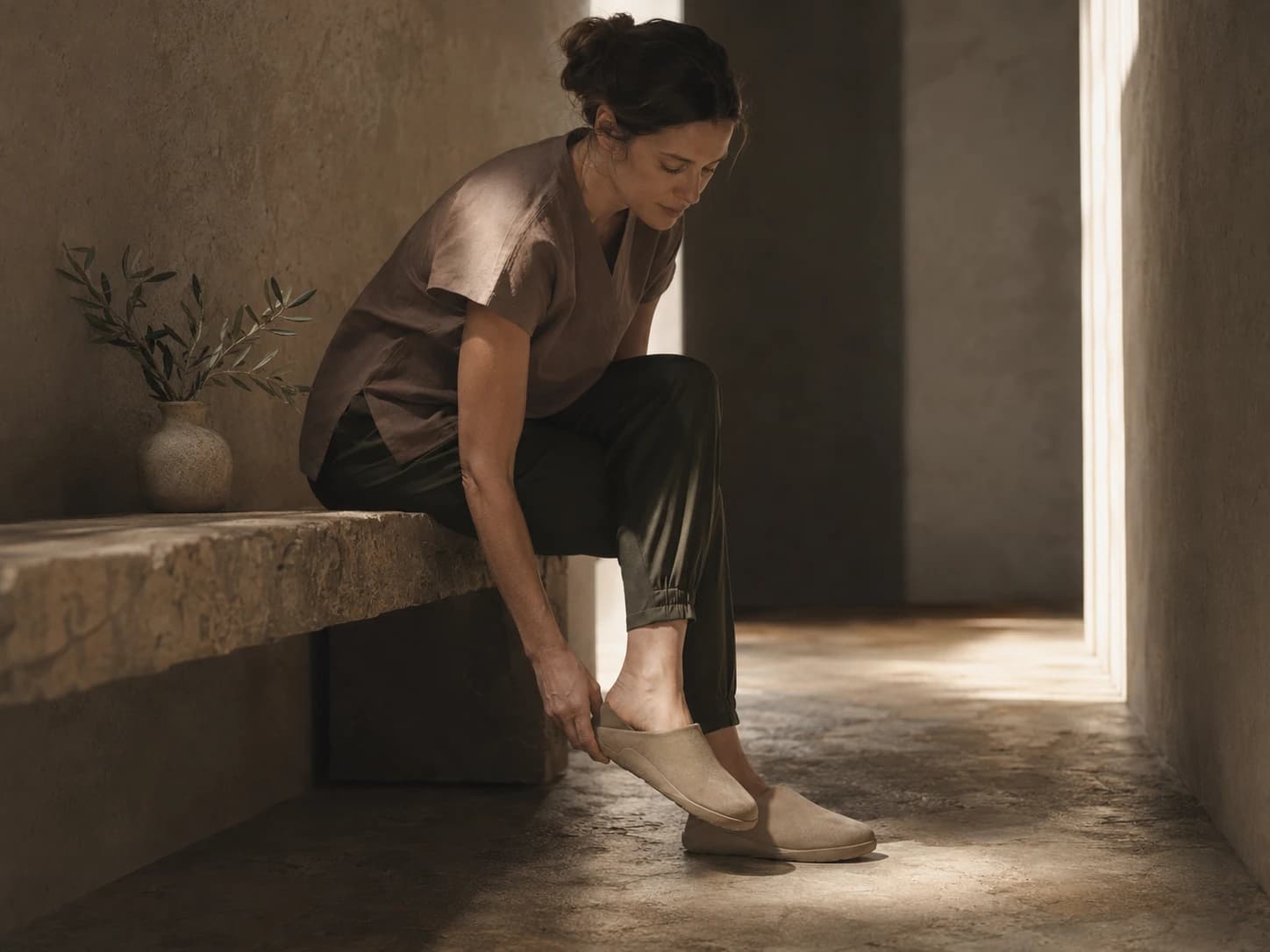 A healthcare worker in scrubs sits on a stone bench and slips on a pair of Inyo mules in warm directional light.