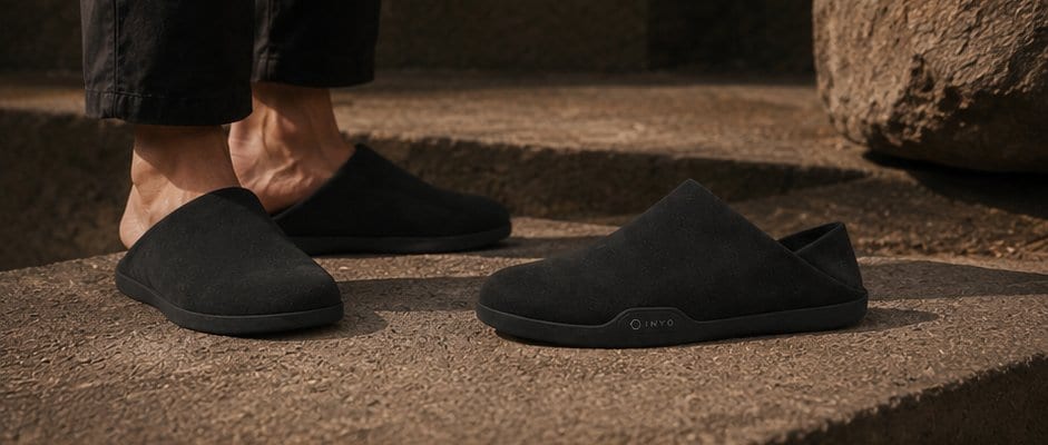 Two black suede mules resting on warm stone beside standing feet.